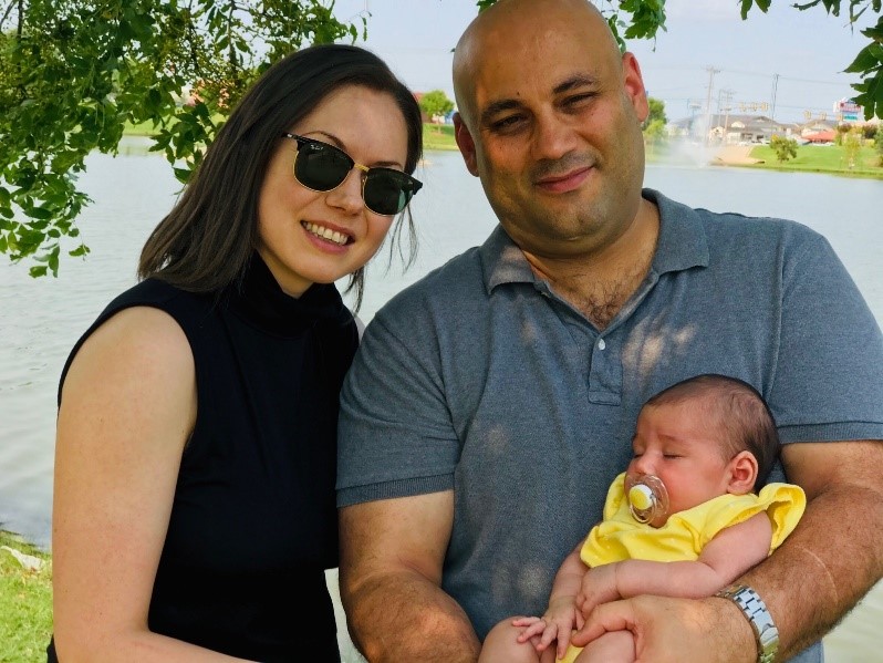 Romina stands next to her husband, who is holding their sleeping baby in his arms. They are both looking to camera and smiling. 